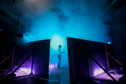 Dunkler Industrieraum der blau beleuchtet ist. In der Mitte stehen Raumtrenner, dahinter eine Person mit dem Rücken zur Kamera.