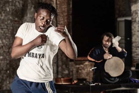 Im Vordergrund steht eine Boxerin mit erhobenen Fäusten. Im Hintergrund sieht man einen Percussionisten auf einer Trommel spielen