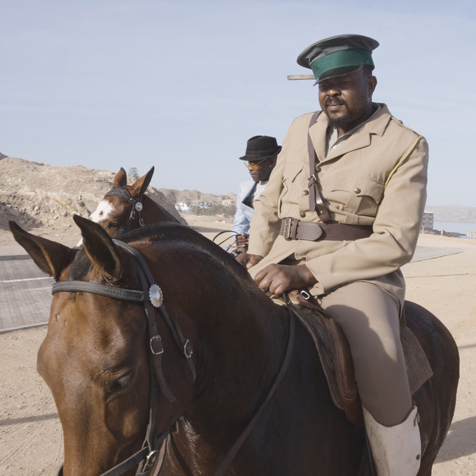 OvaHerero Reiter in deutscher Uniform, der auf einem Pferd durch eine Wüstenladschaft reitet
