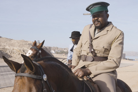 OvaHerero Reiter in deutscher Uniform, der auf einem Pferd durch eine Wüstenladschaft reitet