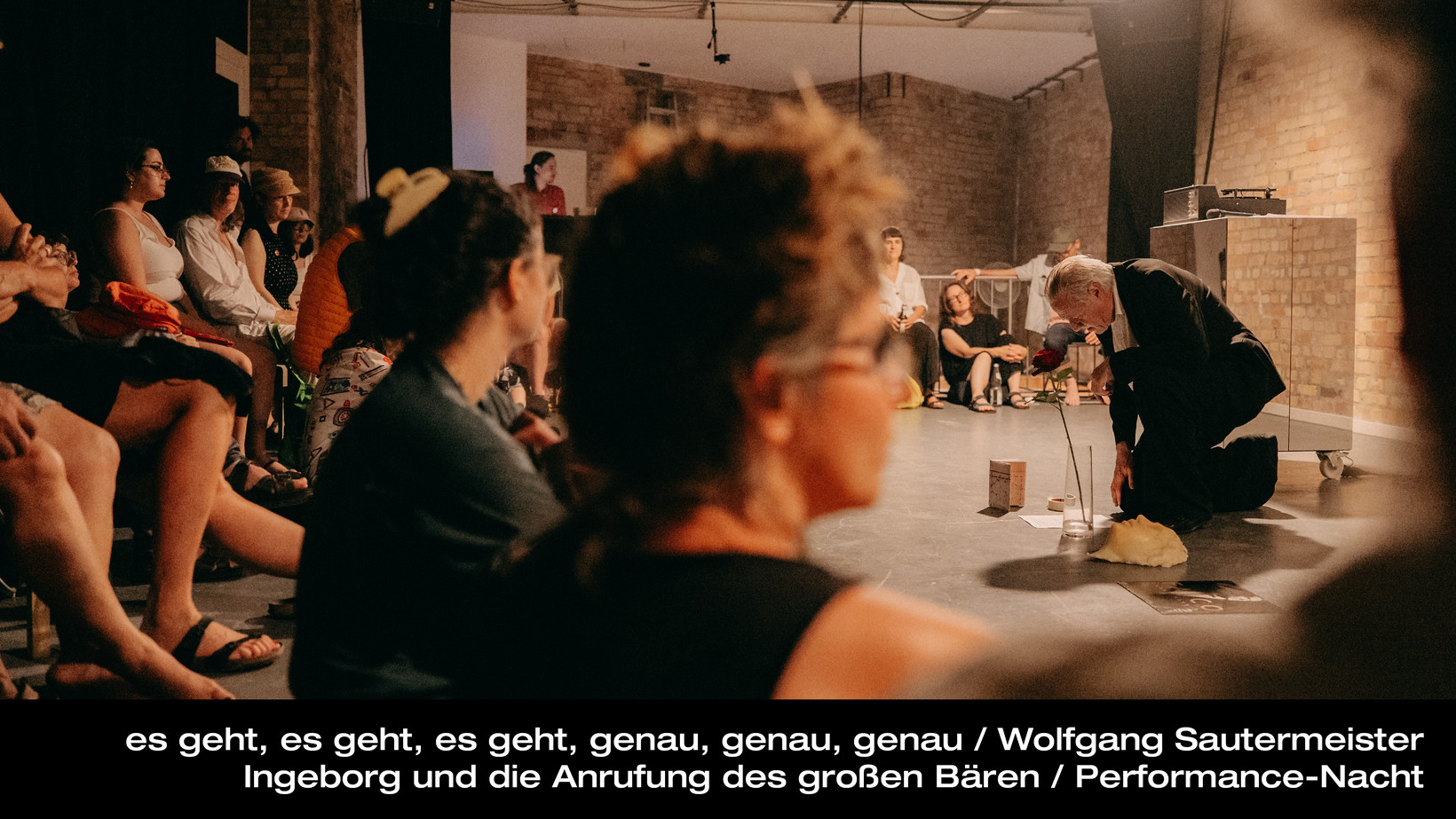 Person kniet auf dem Boden in einem Raum, vor der Person liegen Bilder. Im Hintergrund sieht man Publikum