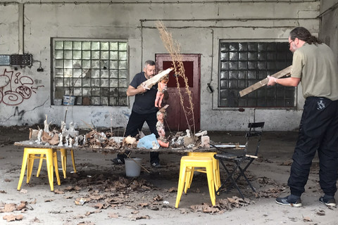 Zwei Personen stehen in einer alten Fabrikhalle. Vor ihnen ein Tisch mit verschiedenem Schrott. Eine Person sägt einer Puppe in den Kopf.