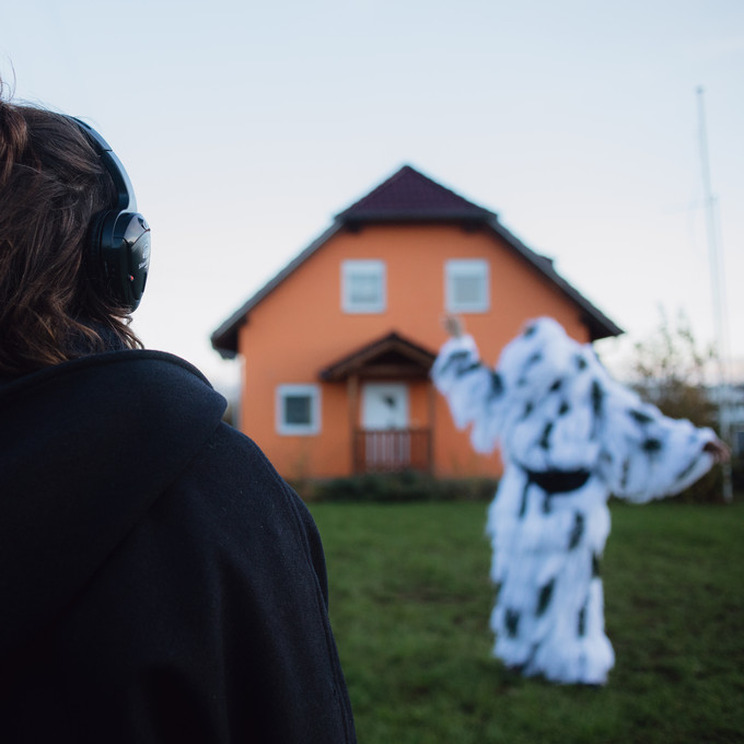 Im Hintergrund ein Haus. Davor eine Figur in einem flauschigen weißen Kostüm mit schwarzen Flecken. Davor mit dem Rücken zur Kamera eine Person mit Kopfhörer. 