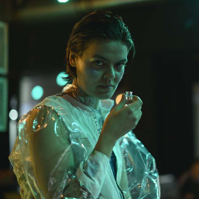 Person in weißem Kittel steht in einem dunklen Raum und schaut auf eine Flasche mit Flüssigkeit in ihrer Hand