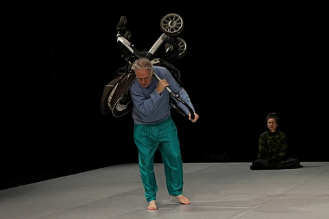 Person läuft mit einem Kinderwagen auf dem Rücken über die Bühne