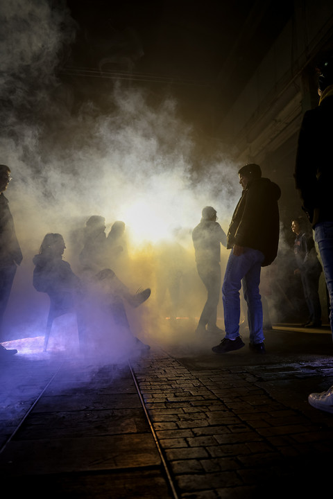 Industrieraum in dem Menschen stehen und sitzen. Durch das Gegenlicht kann man nur Silhouetten erkennen. Der Raum ist verraucht
