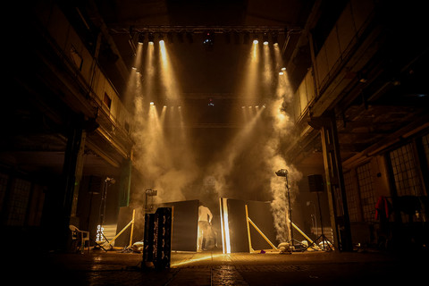Dunkler Industrieraum der gelblich beleuchtet ist. In der Mitte stehen Raumtrenner, dahinter eine Person mit dem Rücken zur Kamera.