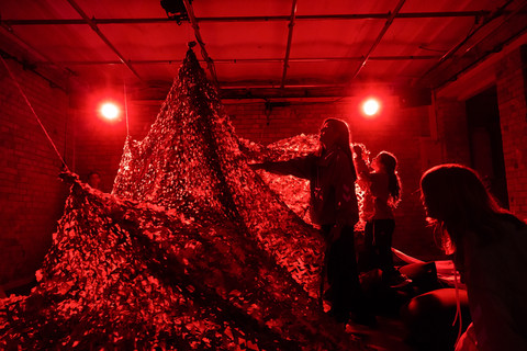 Innenraum mit roter Beleuchtung, darin ein CAmouflage Netz und einige Personen, die man nur schemenhaft erkennt.