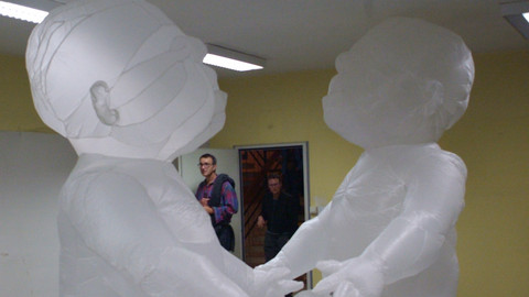 Zwei weiße aufblasbare riesige Babyfiguren, halten sich an den Händen. Sie sitzen mitten in einem Raum.