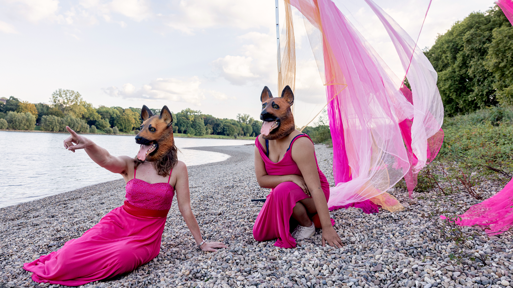 Two persons, wearing pink dresses and dog masks sit on the bank of a river.