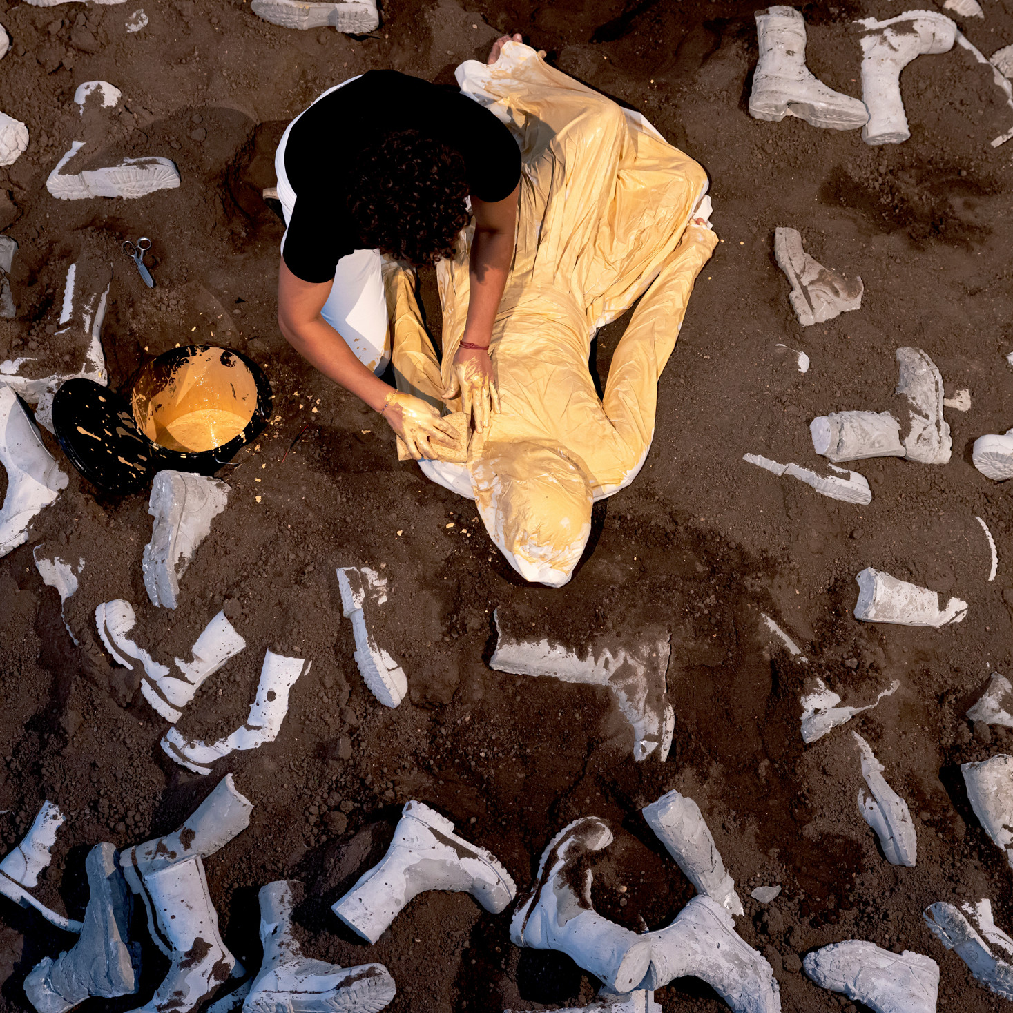 Eine Person bemalt eine andere Person in einem Sack mit goldener Farbe. Sie sitzen auf einem Erdhügel mit Militärstiefeln