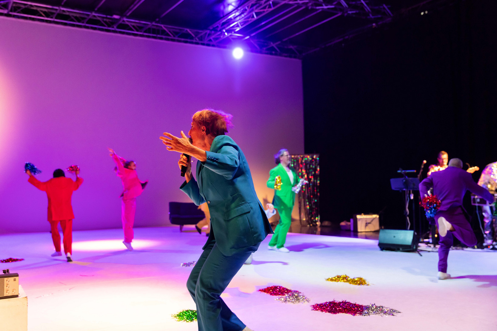 Eine Person im Vordergrund rennt über eine Bühne. Im Hintergrund bewegen sich verschiedene Menschen in bunten Anzügen. 