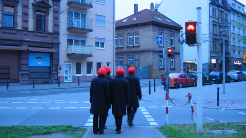 4 Personen in schwarz mit roten Perücken gehen eine Straße entlang. Man sieht sie von hinten