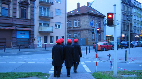 4 people, clad in black walk along a road, wearing red wigs. Seen from behind.