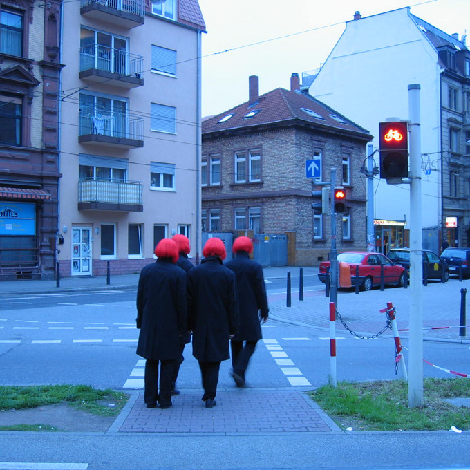 4 schwarz gekleidete Personen mit roten Perücken laufen eine Straße entlang