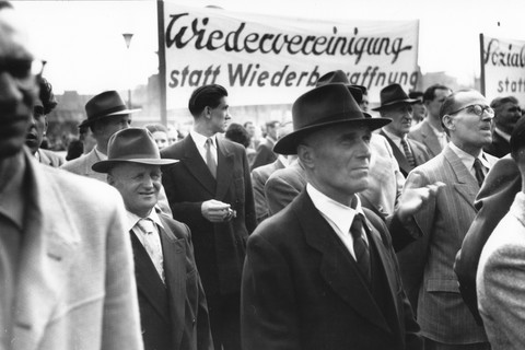 Historisches Bild von Menschen bei einer Demonstration mit einem Schild, das Wiedervereinigung statt Wiederbewaffnung fordert