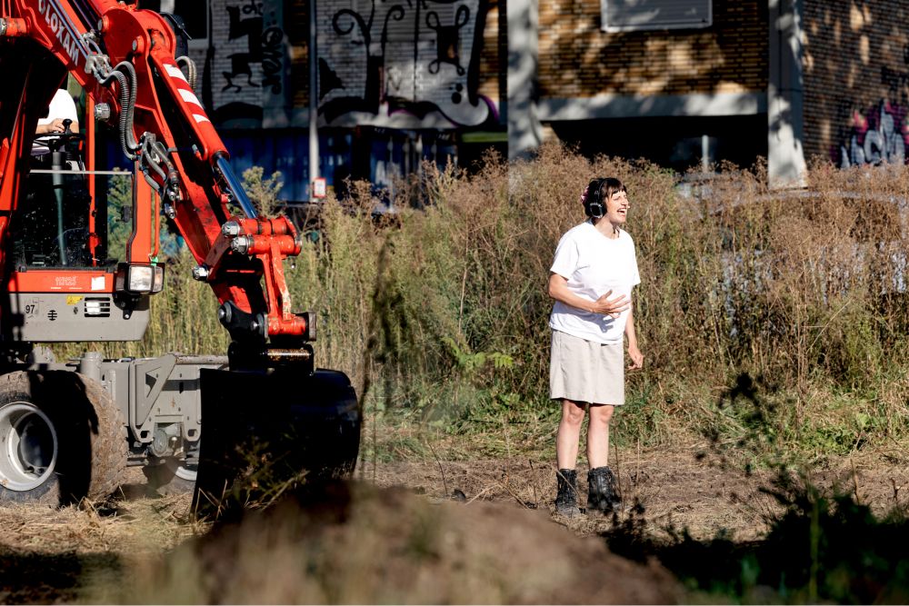 Eine Frau mit Kopfhörer steht auf einer Brachlandschaft vor einem Bagger und spricht