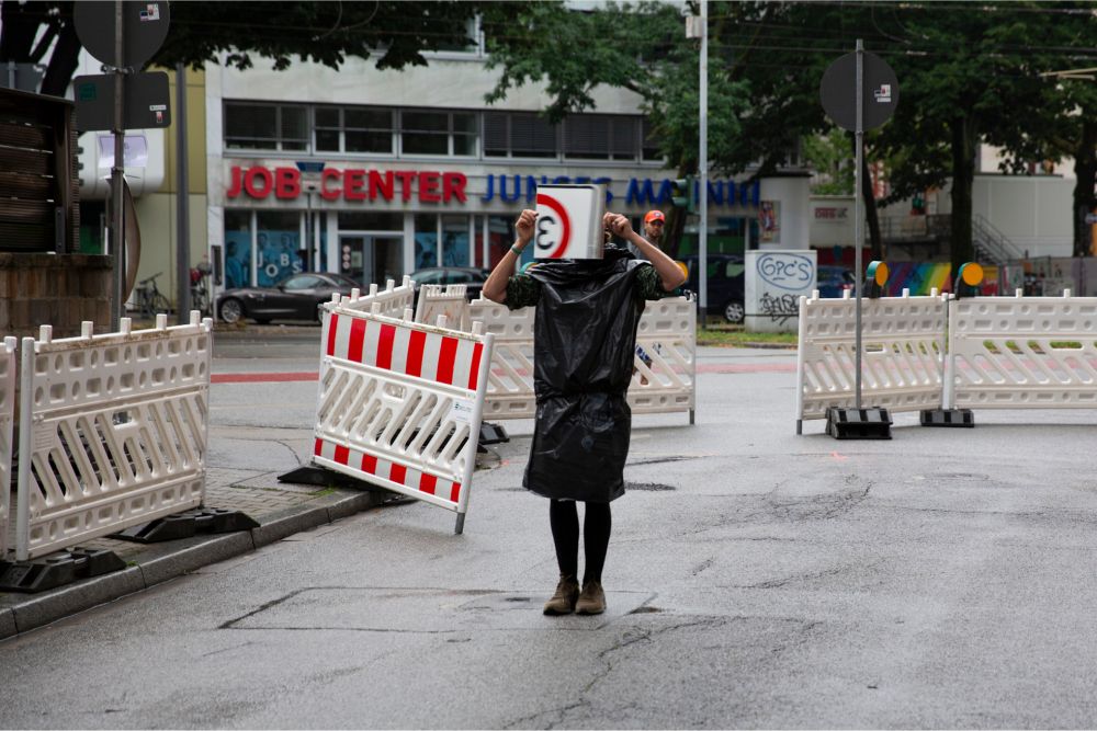 Person steht auf der Straße vor einer Baustelle und hält sich ein Zone dreißig Schild vor das Gesicht.