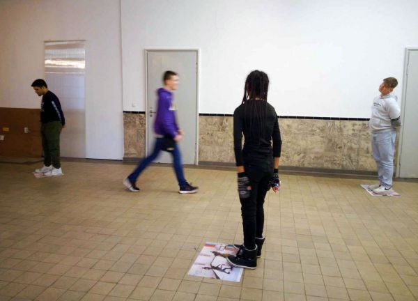 Vier Jugendliche bewegen sich in einem Raum mit gekacheltem Boden und weißer Wand. Im Vordergrund steht ein Mädchen mit dem Rücken zum Betrachter