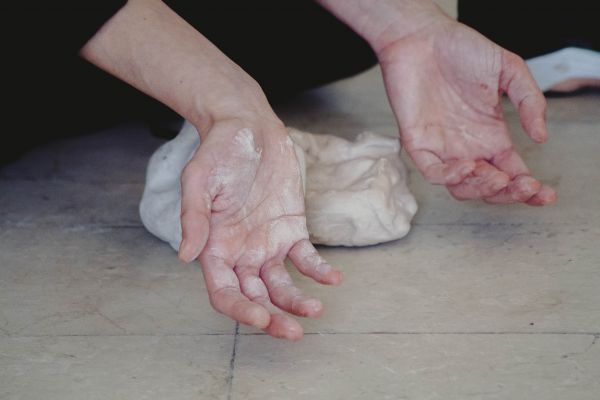 Zwei Hände zeigen mit den Handflächen nach oben in die Kamera. Sie sind mit weißem Ton verschmiert. Im Hintergrund liegt ein Tonklumpen auf dem Boden.