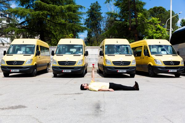 Mann in gelbem T-Shirt und schwarzer Hose liegt vor gelb-schwarzen Vans auf der Straße und streckt einen Arm mit roter Blume in die Luft