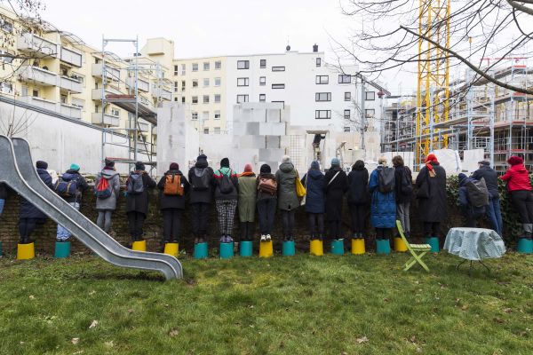 Viele Menschen stehen aufgereiht auf Eimern auf einer grünen Wiese mit dem Rücken zum Betrachter. Vor ihnen steht eine eiserne Rutsche und ein runder Tisch mit karierter Tischdecke. Die Menschen schauen über eine Mauer in eine Baustelle.